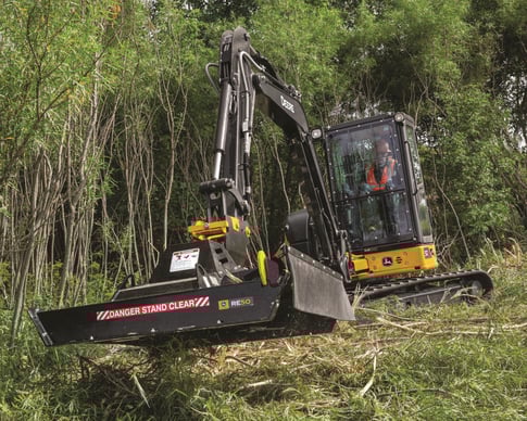 A John Deere 50P excavator is used by a worker to clear brush.