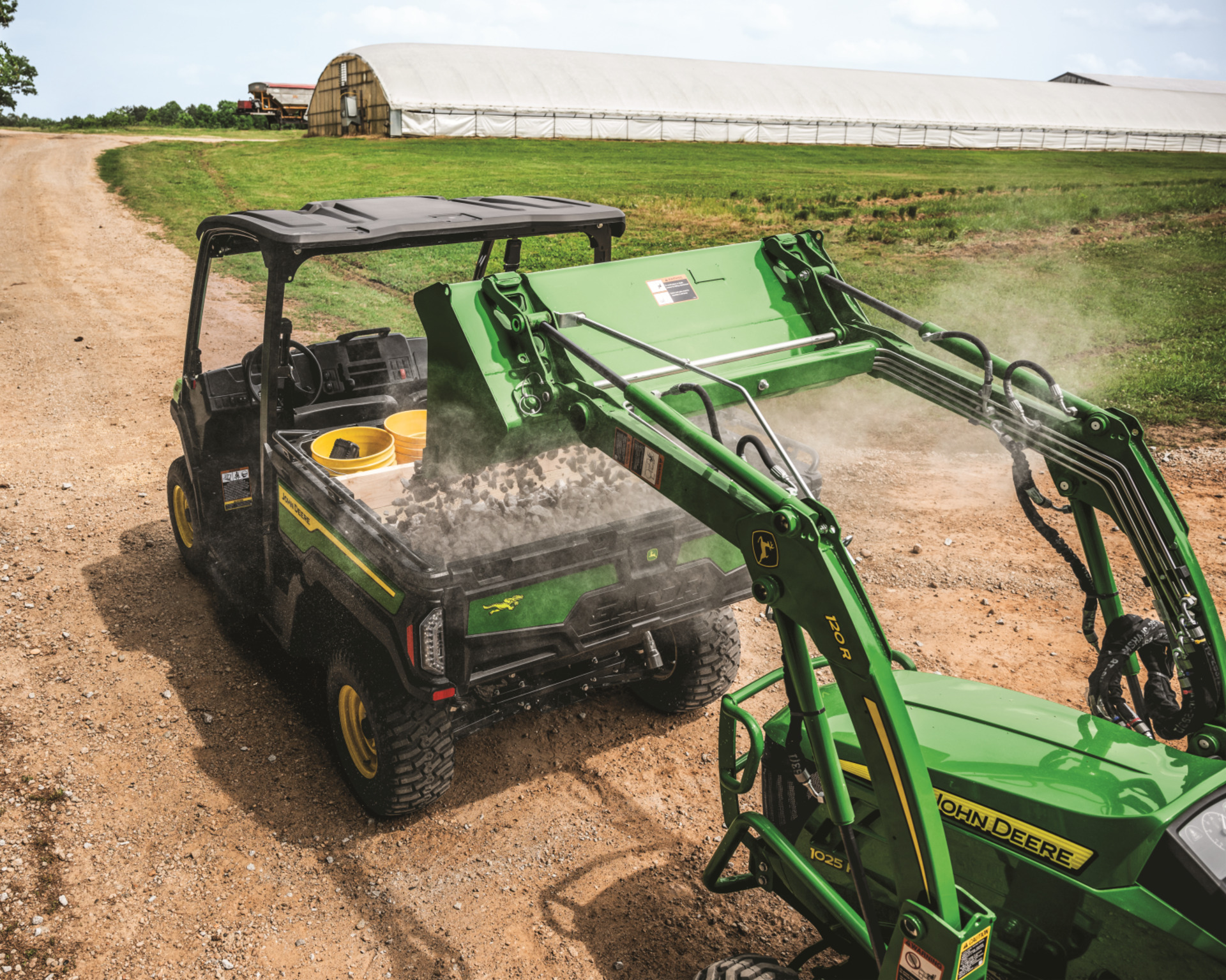 A John Deere Gator gets loaded with gravel by a compact tractor.