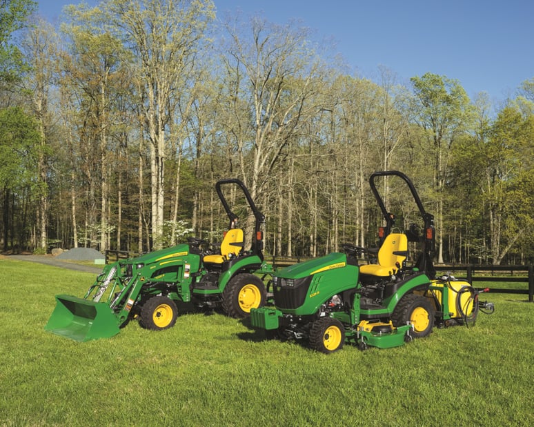 Two John Deere compact tractors with attachments sit in a field.