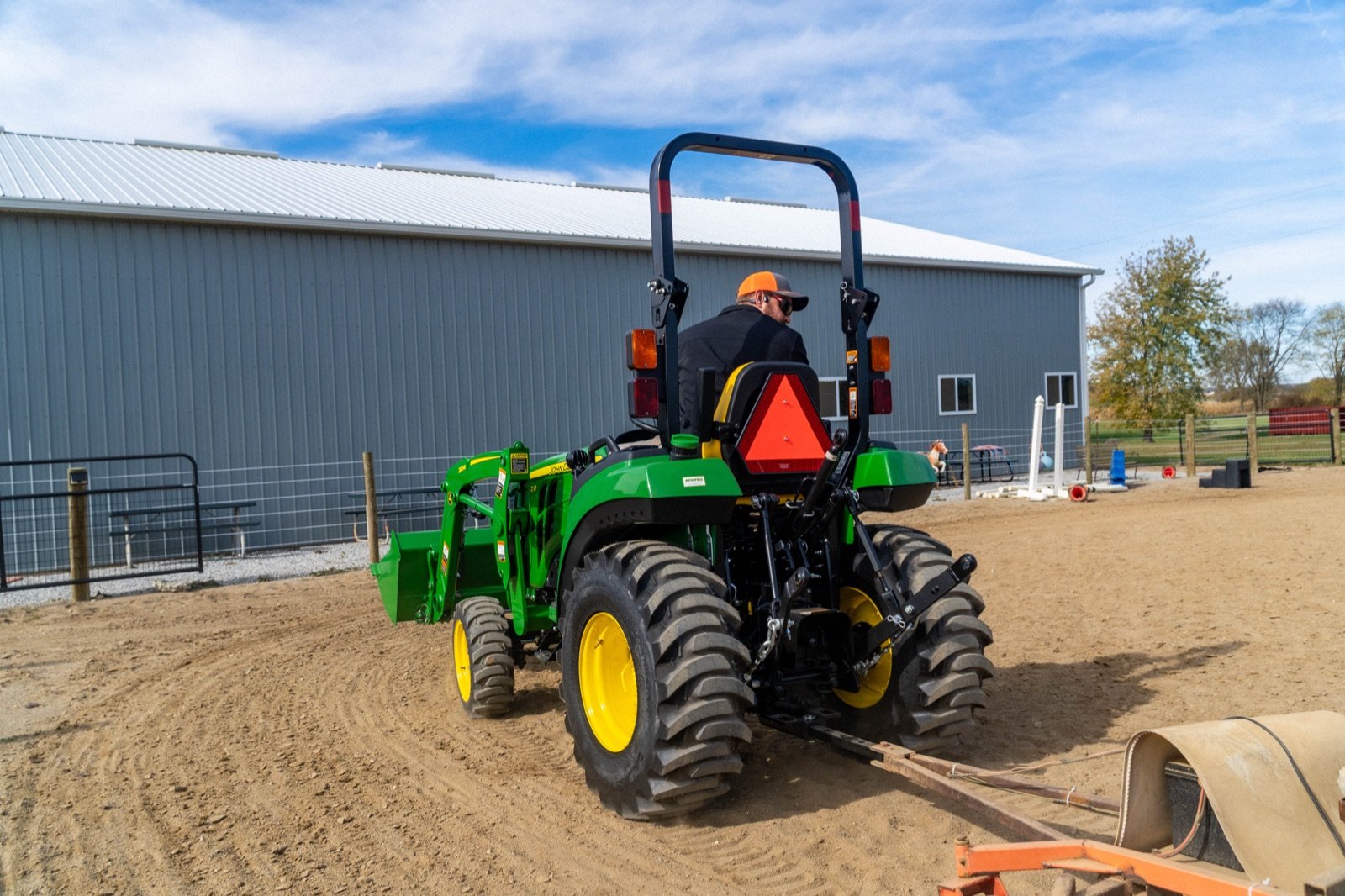 A person drives a green John Deere compact utility tractor with a front loader, pulling an implement on a dirt surface at a r