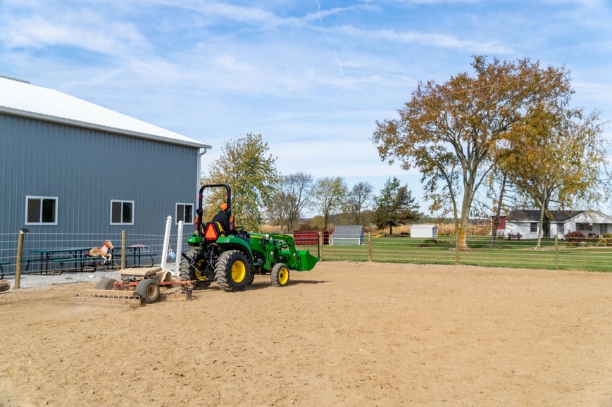 A green John Deere tractor pulls an arena groomer across a dirt field on a farm in autumn.