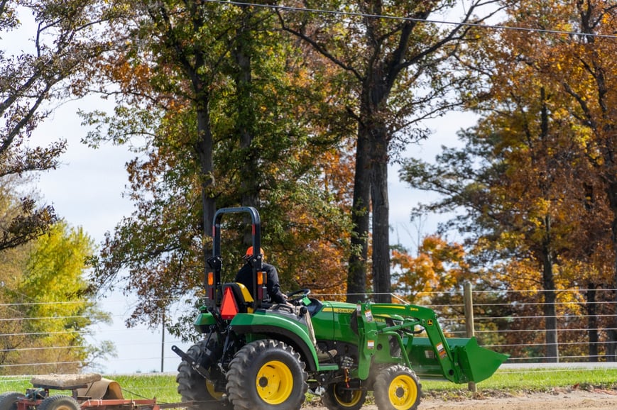 A person drives a green John Deere compact utility tractor with a front loader, pulling an implement on a dirt path, surround