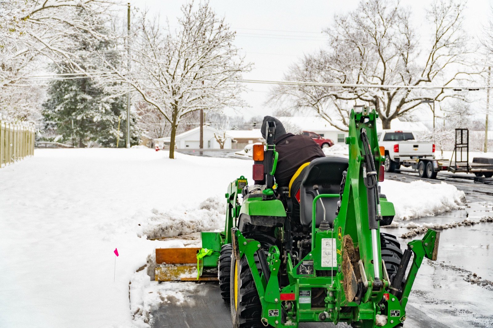 A person operates a green John Deere tractor with a front blade to clear snow from a driveway.
