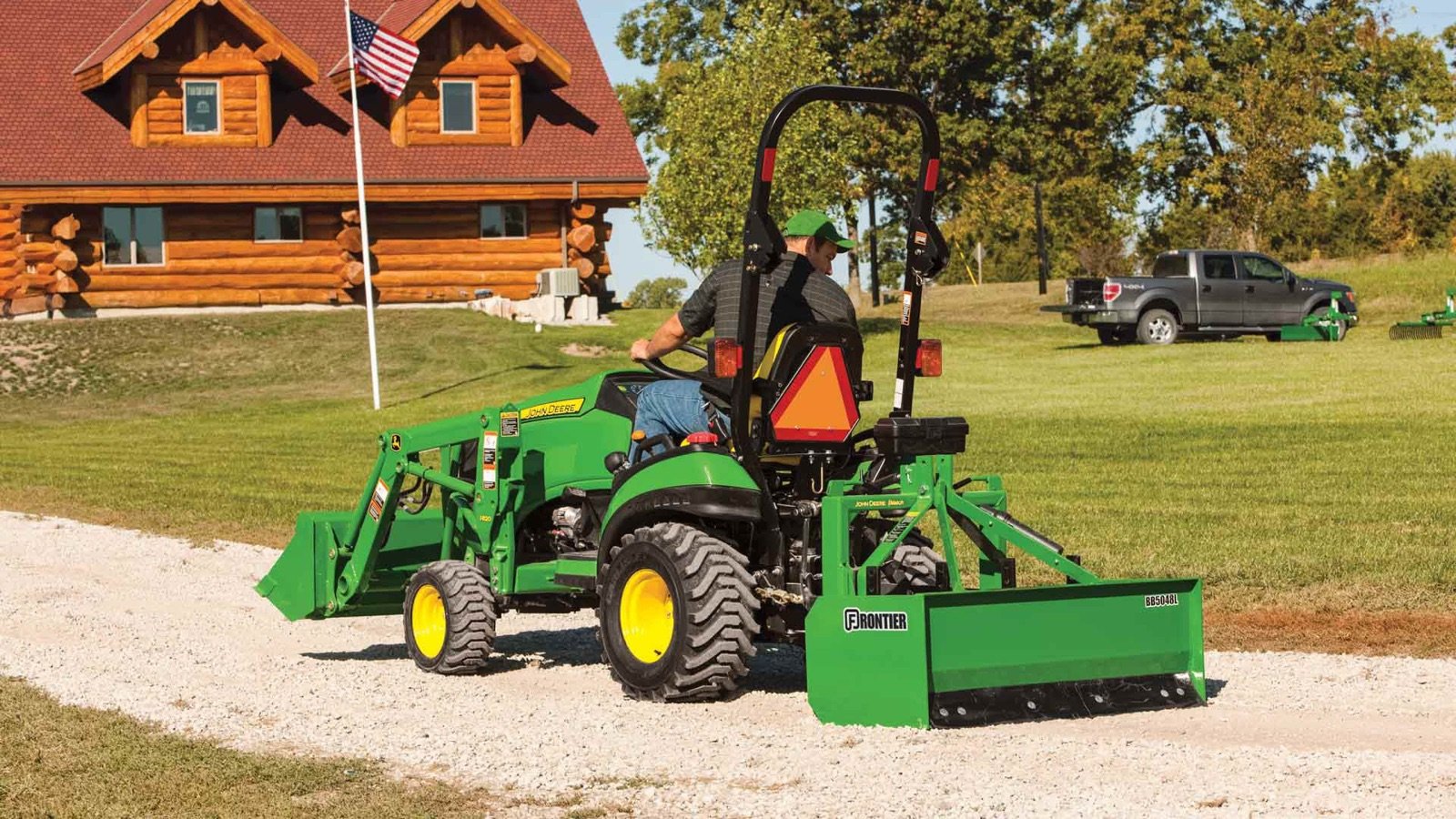 A John Deere compact tractor with a rear box blade is grading a gravel driveway in front of a log cabin.