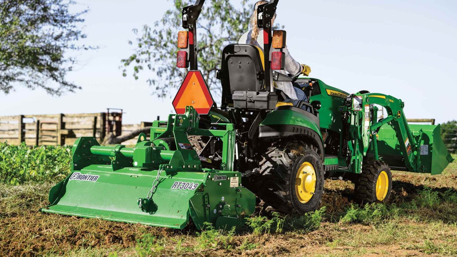 A person drives a green John Deere tractor with a front loader and a rear tiller, tilling soil in a field.