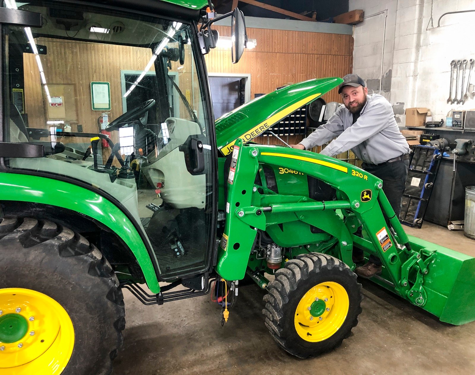 A bearded technician leans over the open hood of a John Deere compact tractor in a service bay.