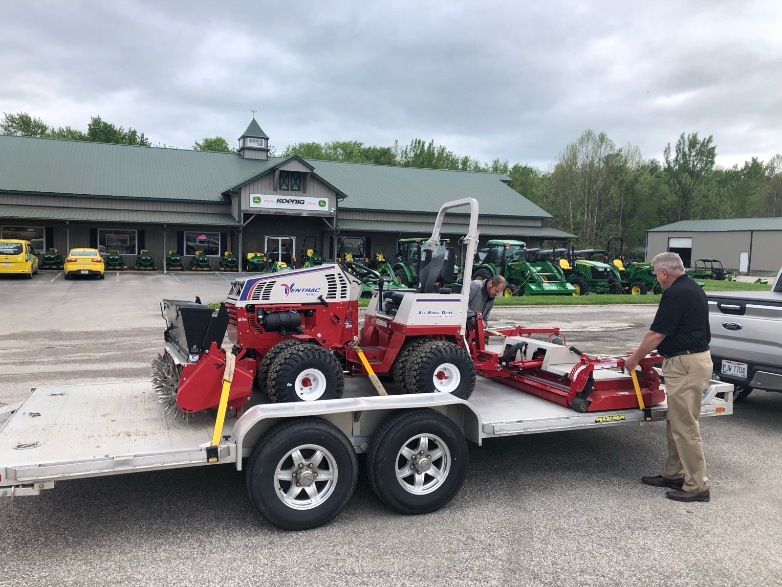 Two men prepare a red and white Ventrac tractor and a mowing deck on a trailer outside a Koenig dealership.