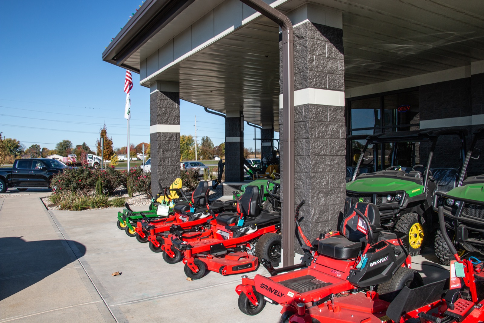 A row of red Gravely zero-turn mowers and green John Deere utility vehicles are displayed outside a dealership on a sunny day