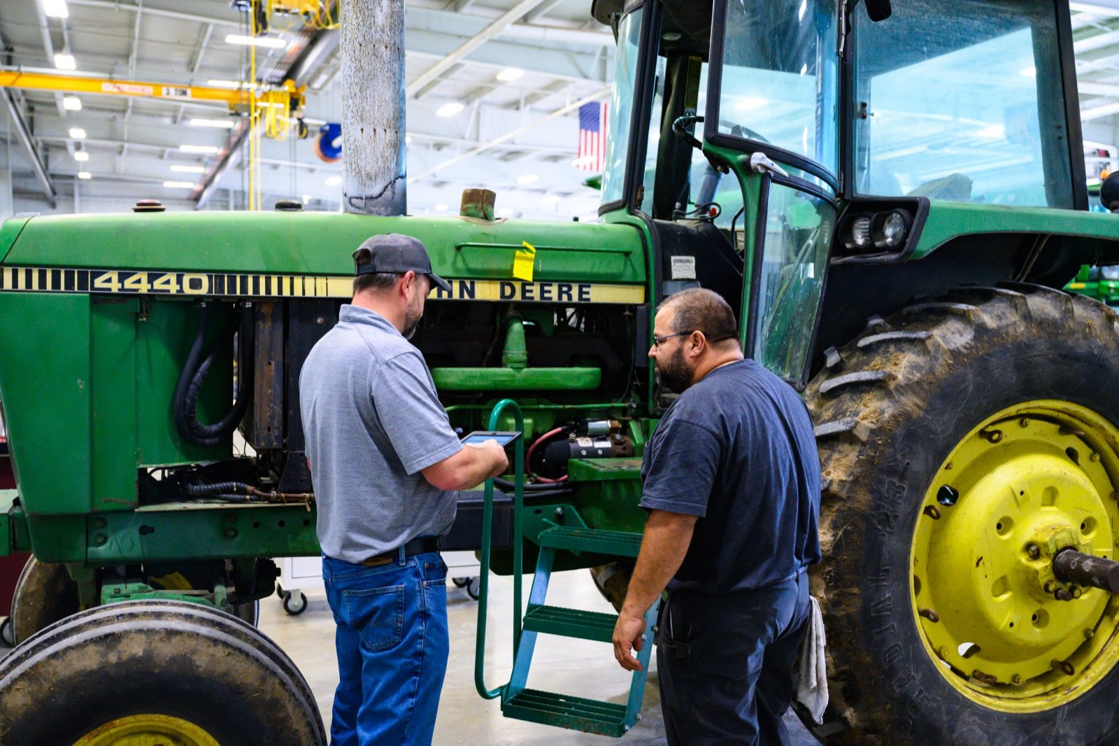 Two technicians inspect a green John Deere 4440 tractor in a service shop, one holding a tablet.