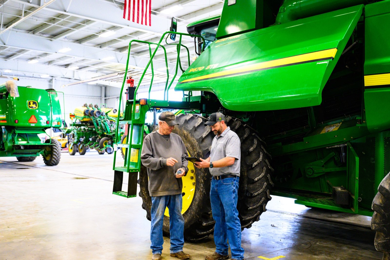 An older farmer and a service writer review a tablet next to a John Deere combine in a dealership.