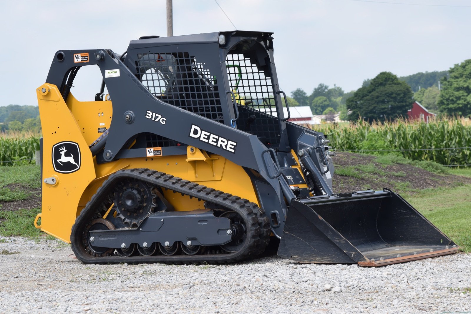 John Deere 317G compact track loader with a bucket parked on a gravel path near a cornfield and farm buildings.