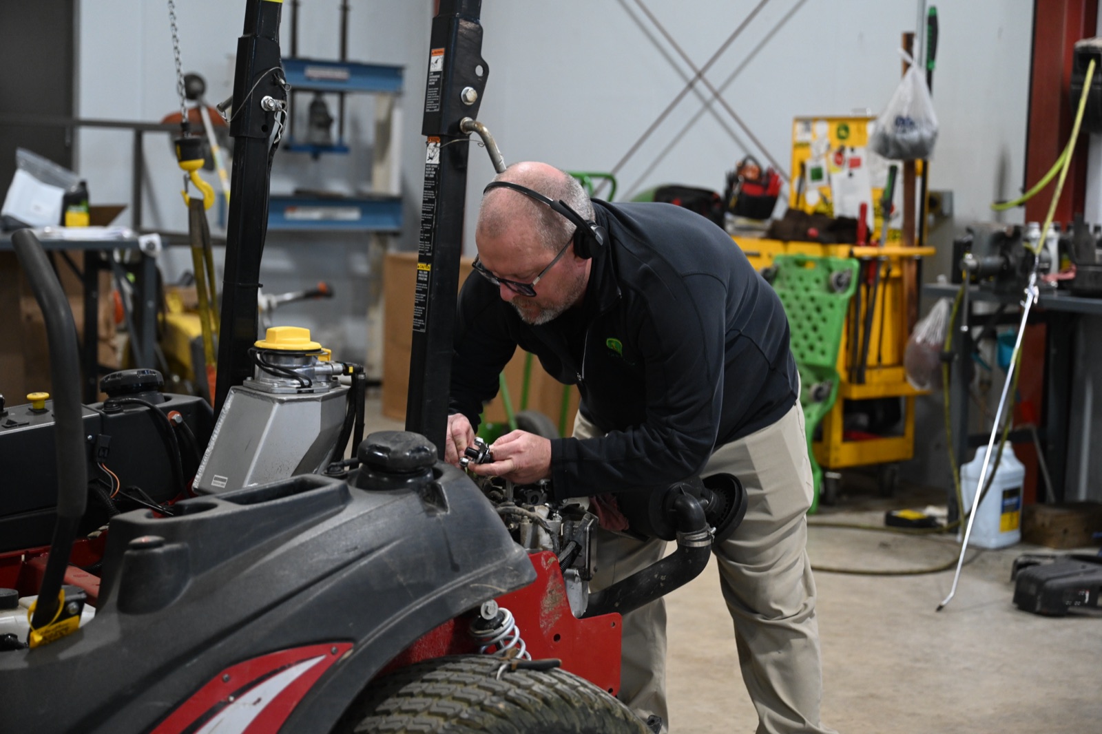 A technician wearing headphones and glasses inspects the engine of a red and black zero-turn mower in a repair shop.