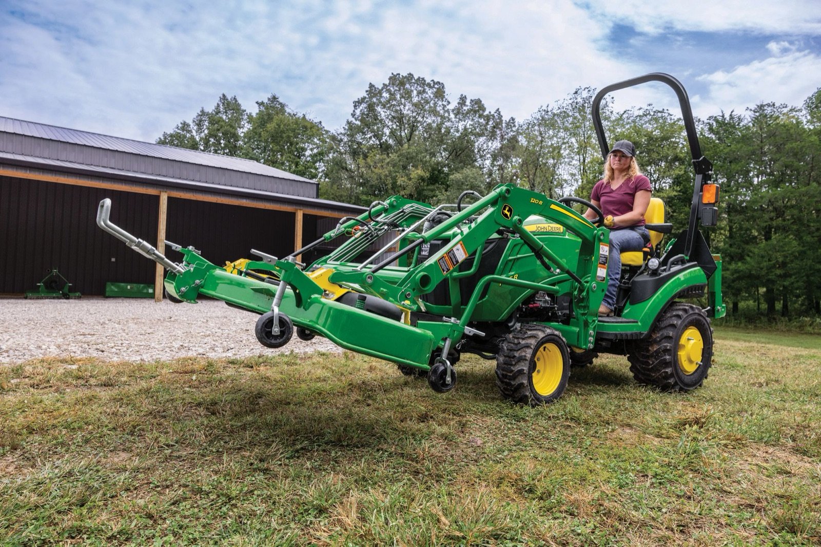 A woman drives a green John Deere compact utility tractor with a front loader and mower attachment near a dark barn.