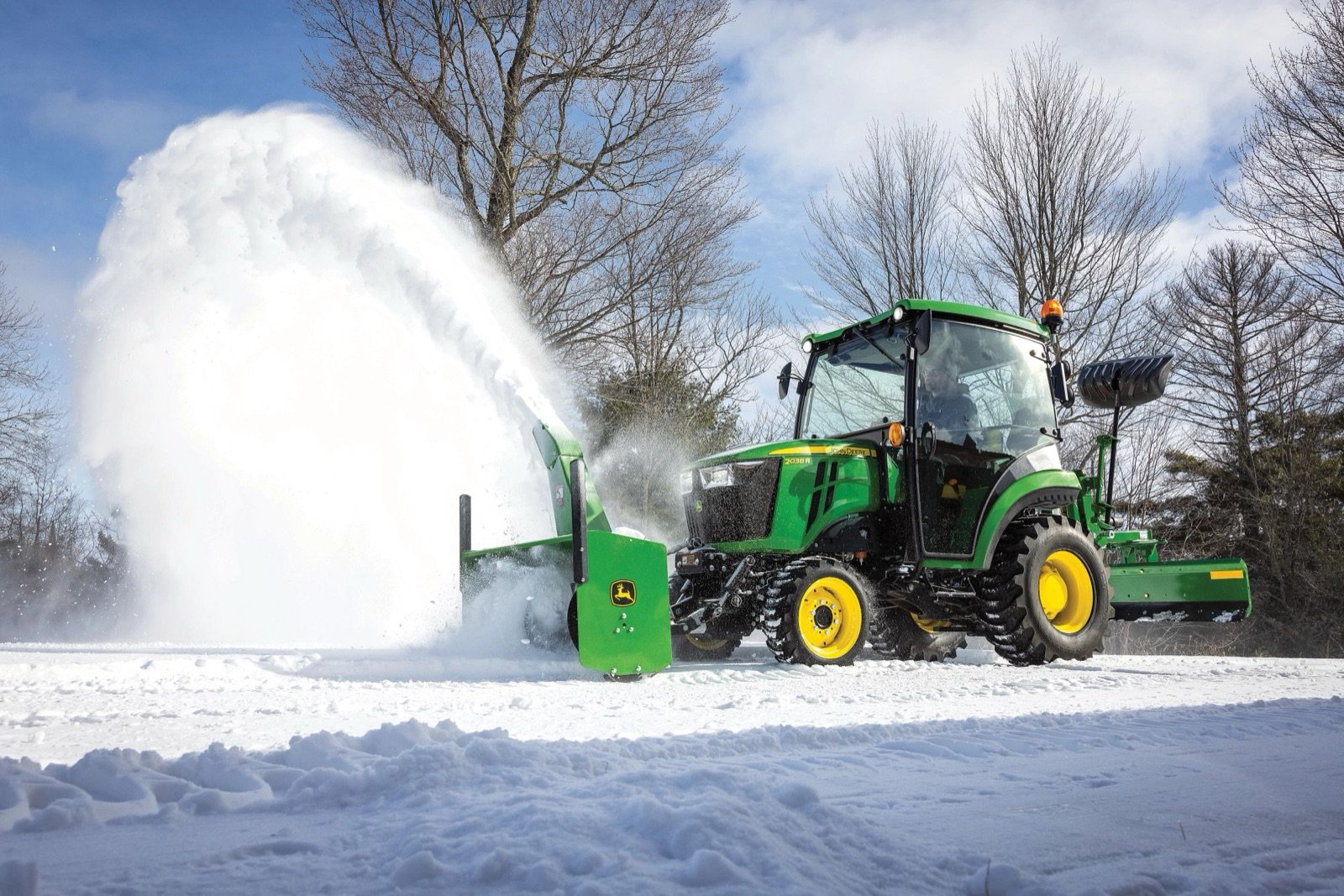 A John Deere compact tractor with a front snow blower actively clears snow on a winter day.