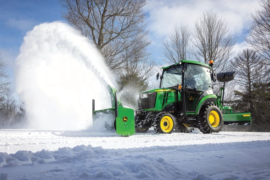 A John Deere compact tractor with a front snow blower actively clears snow on a winter day.