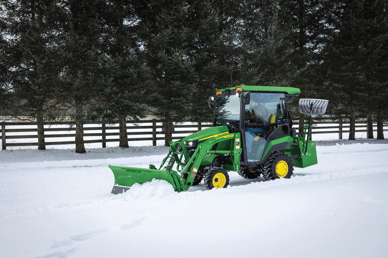 A green John Deere 1025R compact tractor with a front blade clears snow from a driveway lined with trees and a fence.